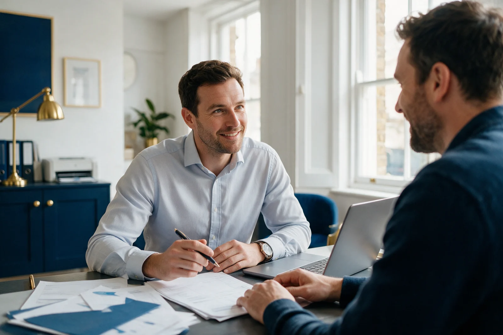 Accountant meeting a self-employed client at a desk in a bright practice office, going through paperwork beside an open laptop