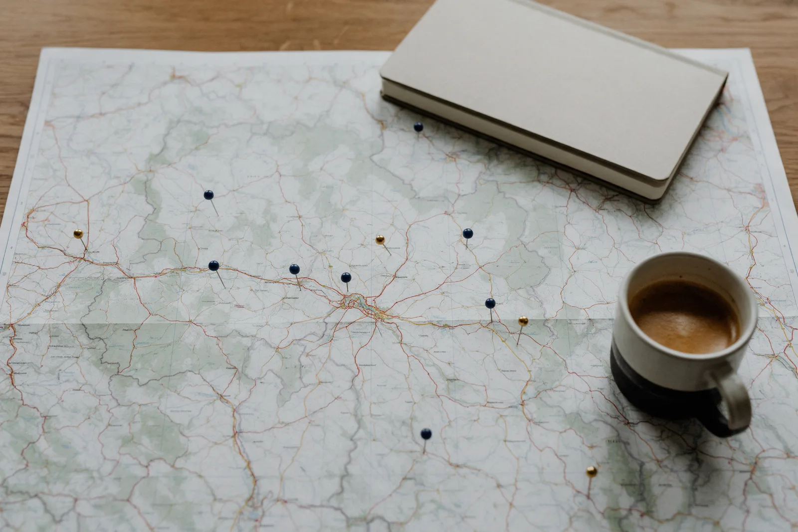 Top down view of a paper regional map on a wooden table with navy and gold map pins, a notebook and a cup of coffee