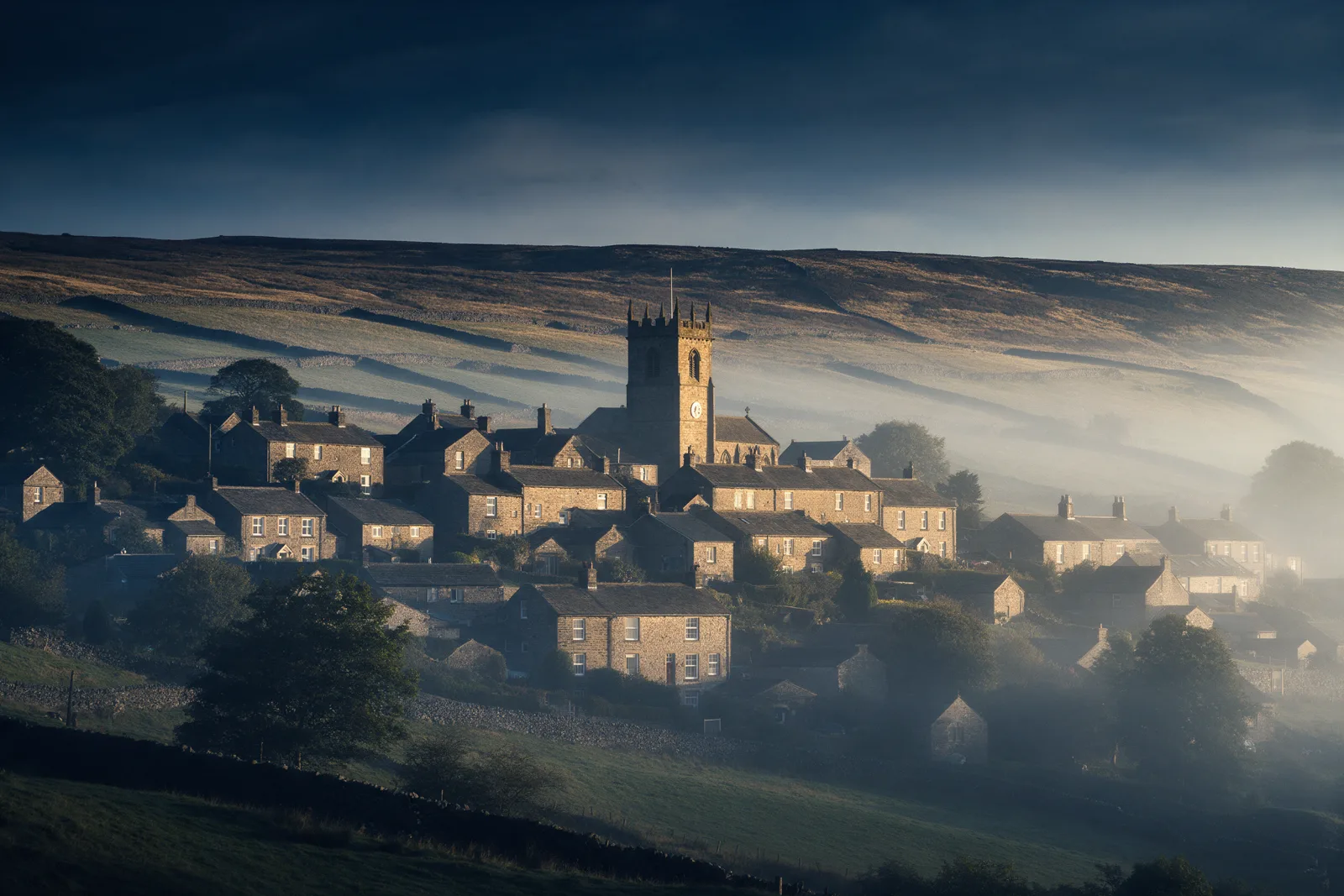 Stone built Pennine village with a church tower on a hillside, dry stone walls and moorland behind on a misty morning
