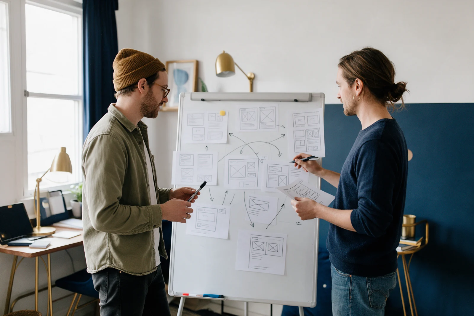 Two creative studio partners planning a new website on a whiteboard of page sketches in a small light-filled office