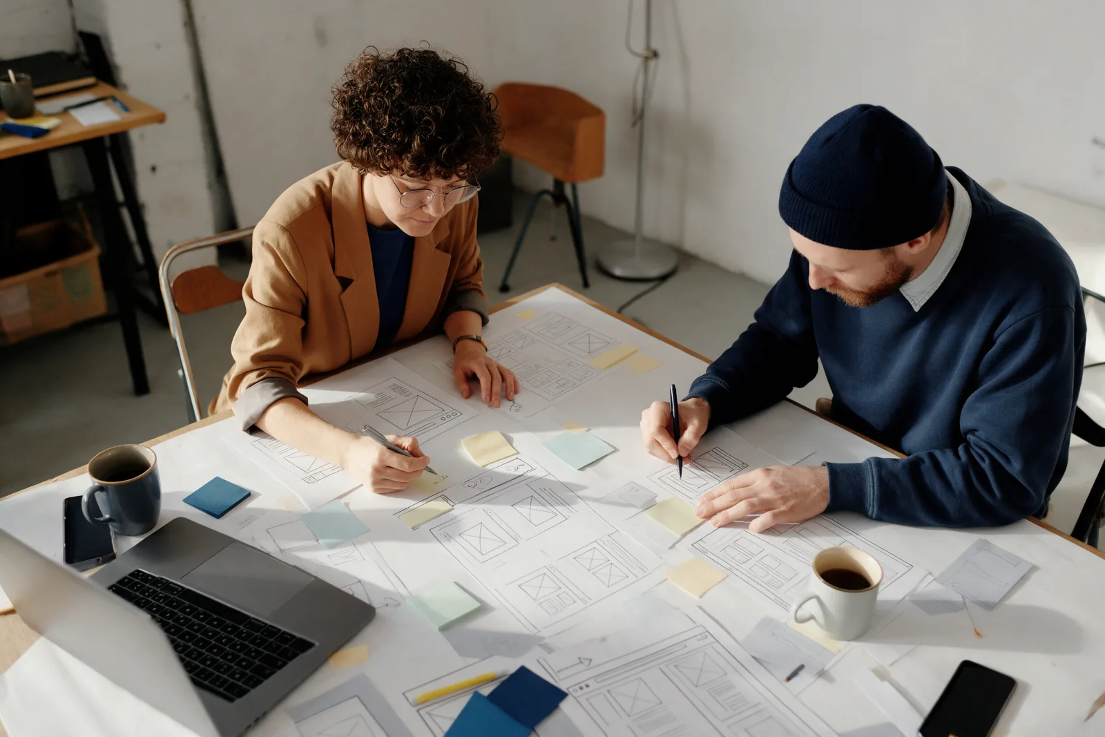 Two designers sketching website wireframes with pens and sticky notes on a large table, photographed from above